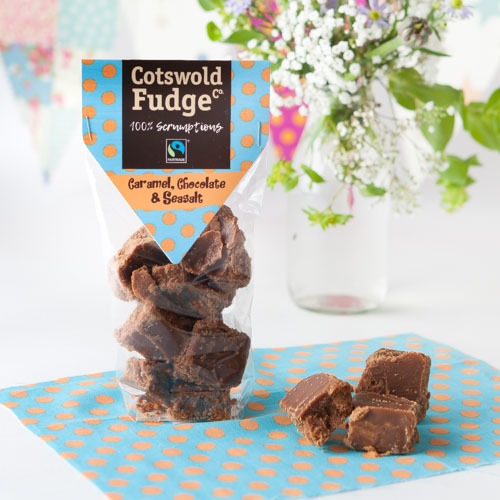 A clear, standing pouch of Cotswold Fudge Co.'s caramel, chocolate, and sea salt fudge is displayed. The package is decorated with a blue and orange polka dot design, with visible chunks of fudge spilling out onto a matching polka-dotted napkin. The fudge is rich brown with layers, suggesting a creamy texture. In the background, a clear vase holds a bouquet of flowers, adding a fresh and inviting atmosphere to the image. The overall setting is bright, with a cheerful, spring-like ambiance.