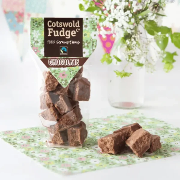 A standing pouch of Cotswold Fudge Co.'s chocolate fudge is displayed, featuring a decorative green and floral patterned design on the packaging. The label reads "Chocolate," and visible chunks of rich, brown chocolate fudge are stacked in the clear package. A few pieces of the fudge are arranged on a matching floral napkin in front of the package. In the background, a clear vase holds a bouquet of delicate white flowers and green leaves, adding a fresh, spring-like touch to the setting. The scene is bright and airy, creating a sweet and inviting atmosphere.