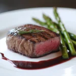 tender fillet steak, cooked to a perfect medium-rare with a slight char on the outside, served on a white plate with asparagus spears and a drizzle of red wine reduction