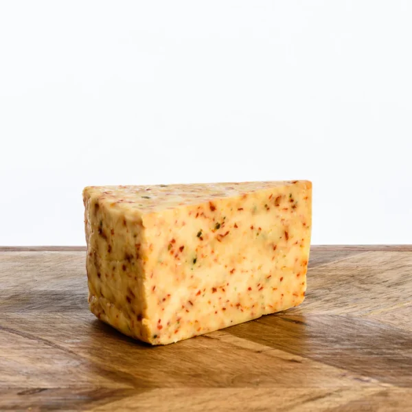 A triangular wedge of artisan Burner cheese infused with vibrant flecks of red chilli and herbs, placed on a wooden chopping board. The cheese has a smooth, slightly waxy texture, highlighted by natural light against a clean white background.