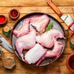 Raw jointed rabbit pieces arranged in a pan, surrounded by spices, herbs, and cooking utensils on a rustic wooden surface, ready for preparation.