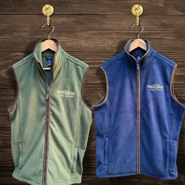 The Black Farmer gilet available in moss green and navy blue, featuring a sleeveless design with an embroidered logo, displayed on wooden hangers.